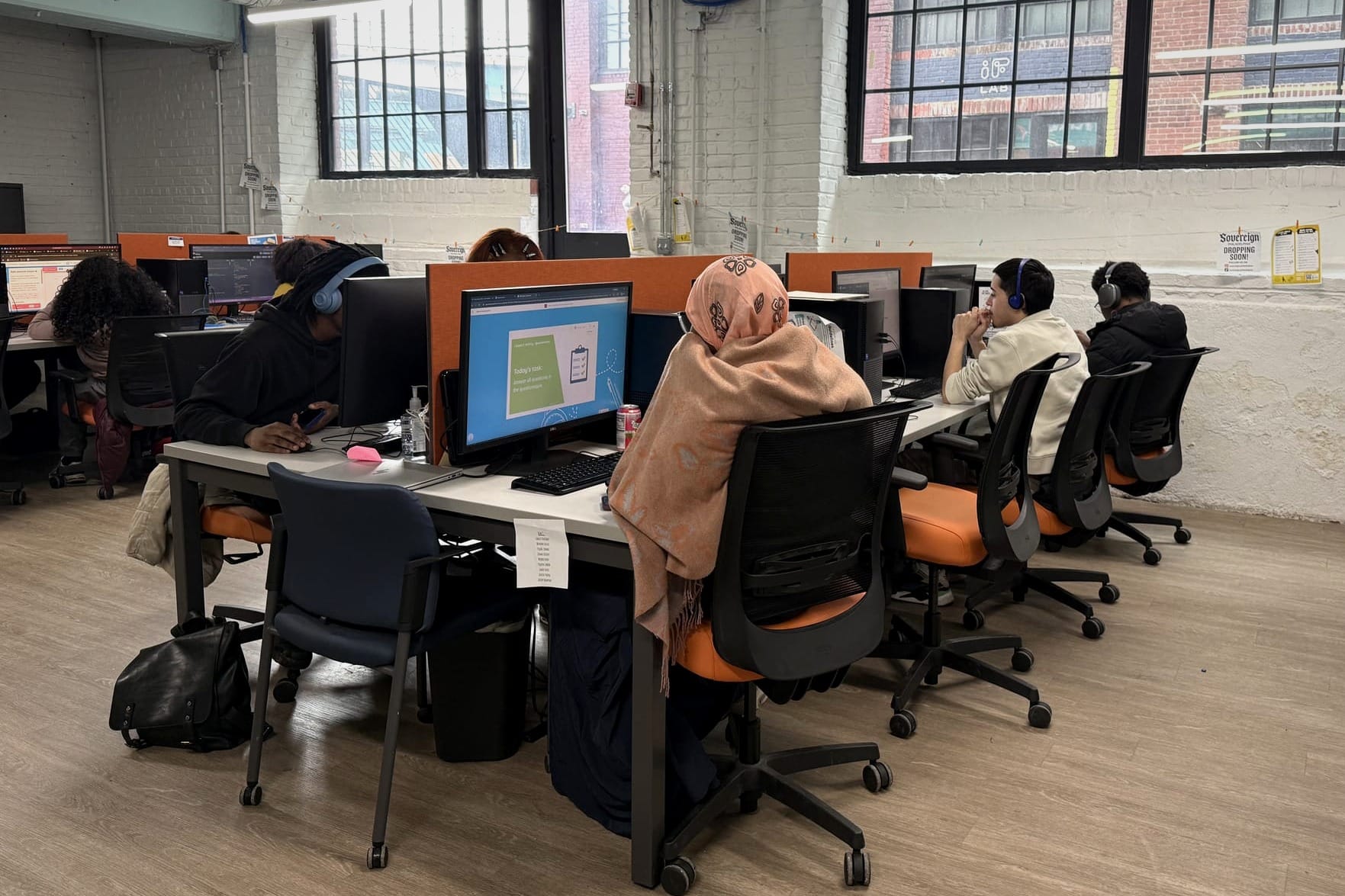 Four young adults wearing headphones work at desktop computers in a bright room with industrial windows and orange chairs.